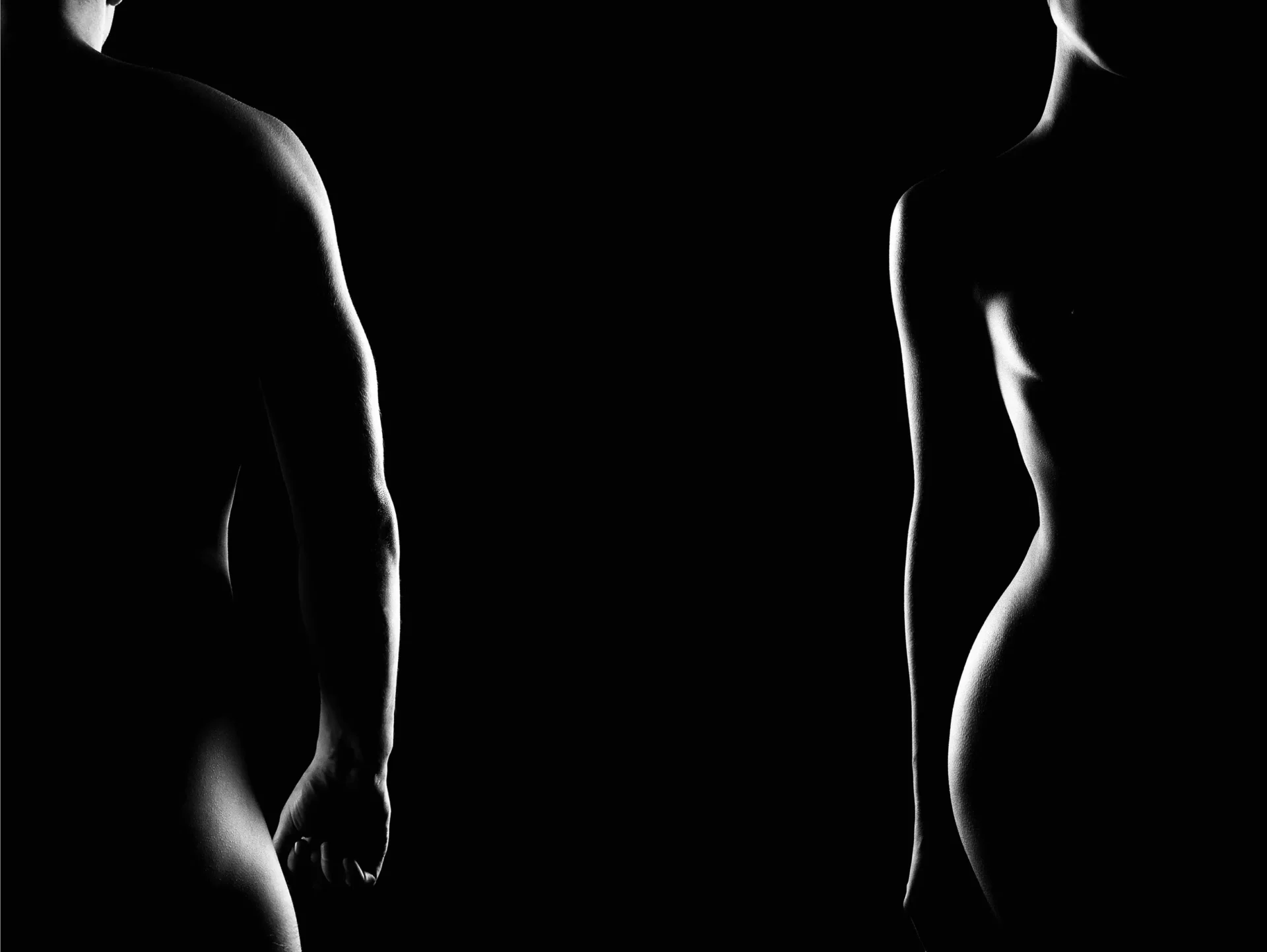 Two naked men standing in the dark with one looking at the other.